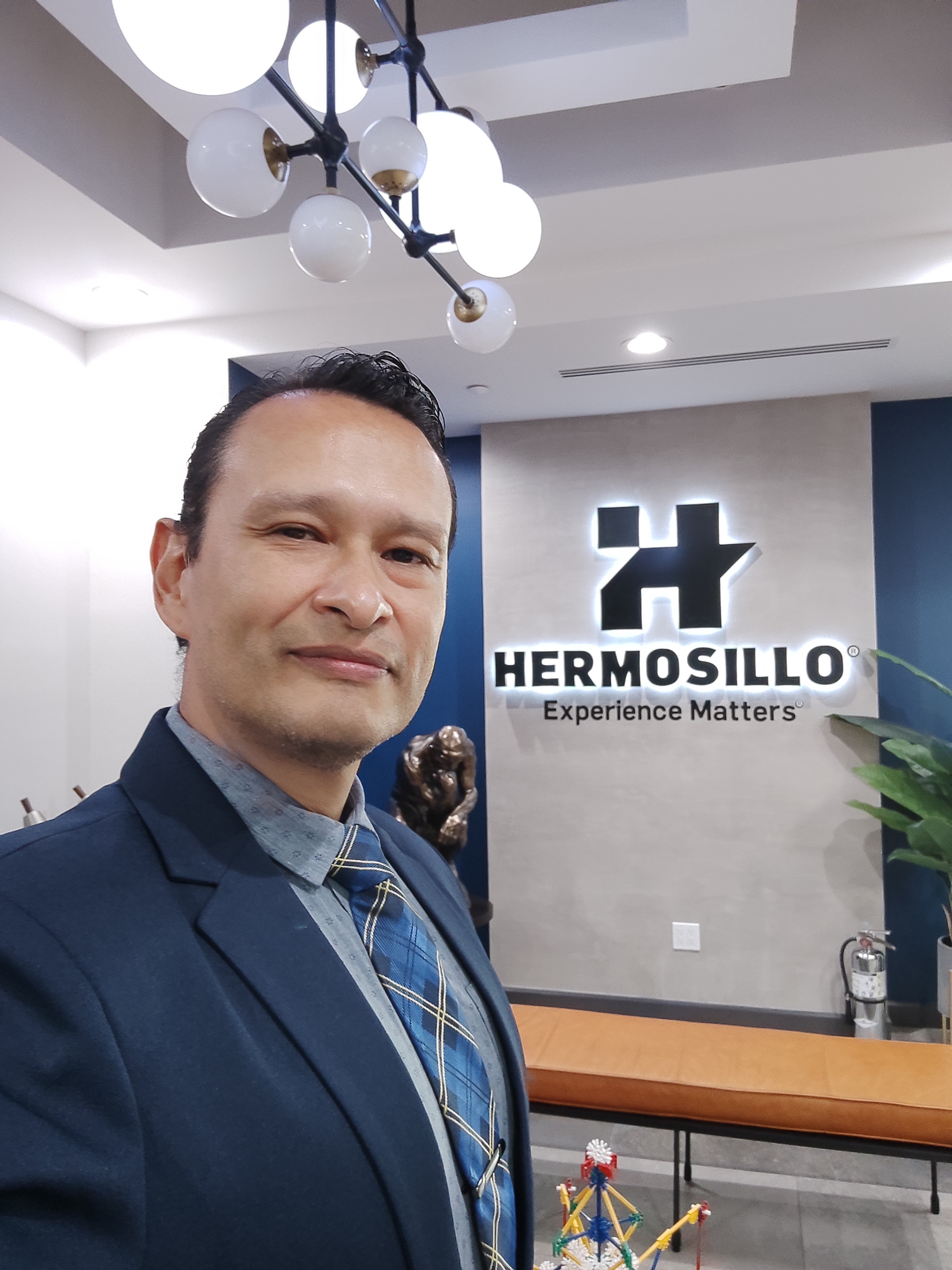 Gustavo frente al logotipo de Grupo Hermosillo, traje azul y corbata azul‑amarilla